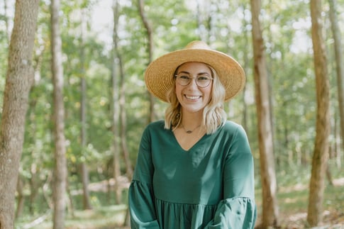 A smiling woman with blonde hair, glasses, and a straw hat stands in a sunlit forest. She is wearing a flowing green dress and looking directly at the camera, surrounded by tall trees and lush greenery.