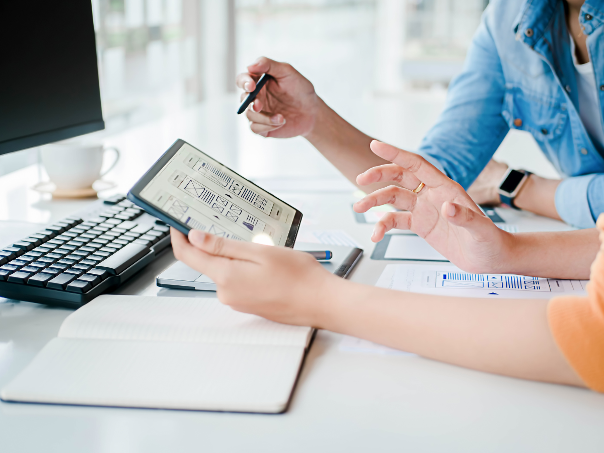 Two people collaborating at a desk, reviewing a tablet displaying wireframe designs. One person gestures while holding a stylus, while the other holds the tablet. A computer keyboard, notebook, and documents with charts are also on the desk, creating a professional and productive workspace