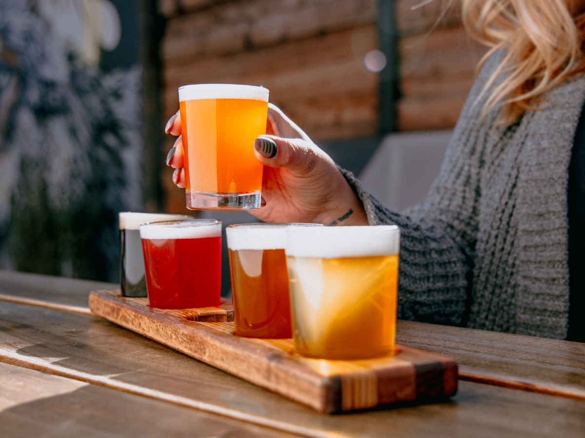 A hand holds a glass of hazy, golden beer from a wooden flight paddle with four craft beers in varying colors, set on a sunlit outdoor table