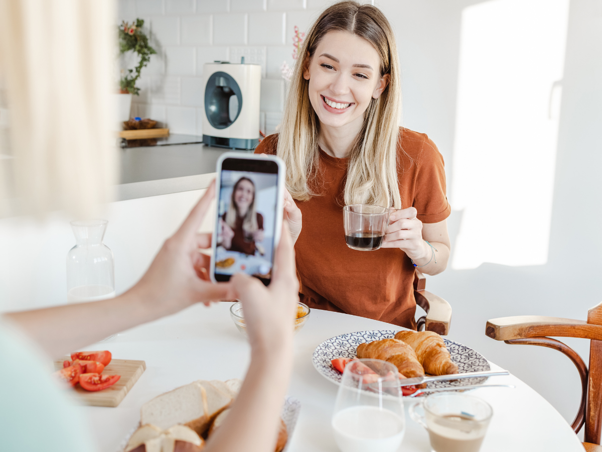 A young woman with long blonde hair and a brown shirt smiles while holding a glass of coffee at a bright, modern breakfast table. In the foreground, another person takes a photo of her with a smartphone, capturing the moment. The table is set with croissants, sliced bread, tomatoes, and drinks, creating a cozy and inviting atmosphere